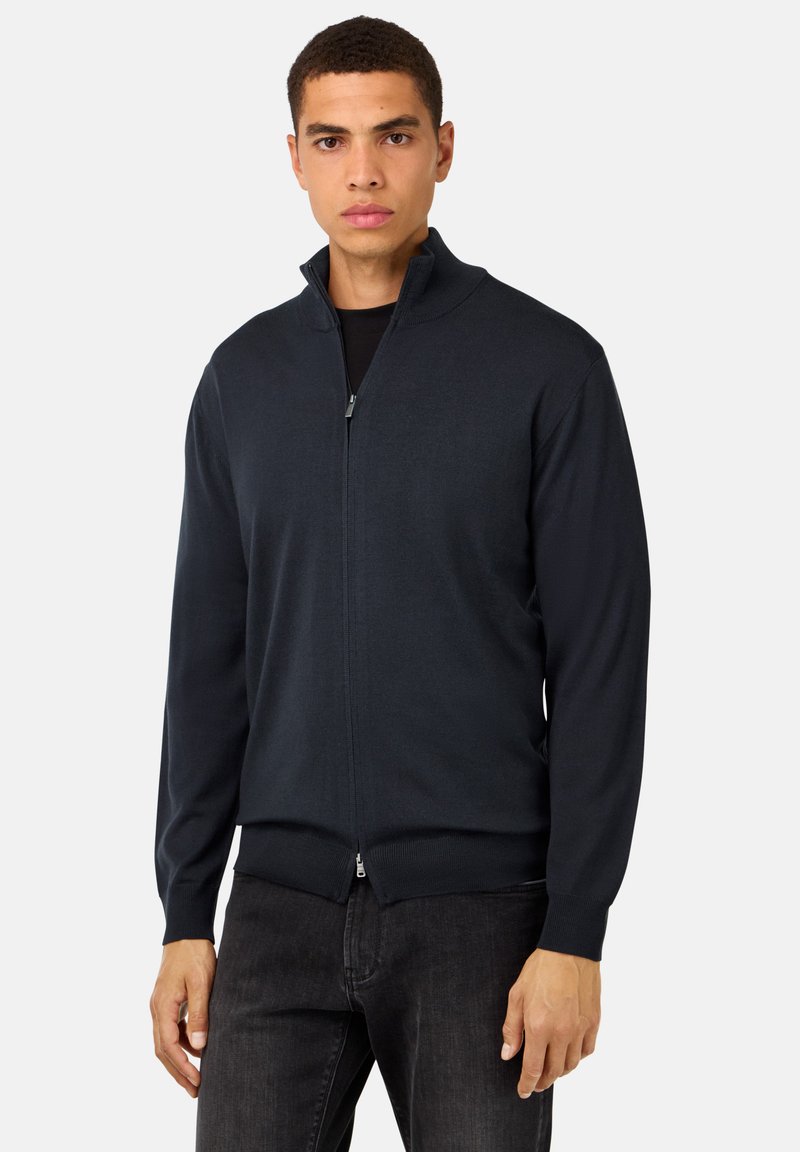 Moška zadrgo zapirana cardigan v temno modri barvi. Pletenina, klasičen kroj, rebrasti manšeti in rob. Zadrga s visokim ovratnikom. Priložnostna zasnova.