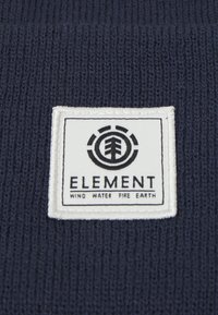 Tejido de punto azul marino con una etiqueta rectangular blanca que presenta un logo negro de Element y el texto: "VIENTO AGUA FUEGO TIERRA."