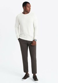 Witte geribde lange mouwen trui, bruine slim-fit broek en zwarte sneakers met witte zolen. Horloge om de linkerpols, handen in de zakken.