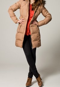 Chaqueta acolchada beige con capucha forrada de piel, cintura ceñida y cremallera frontal. Combinada con jeans negros y botas de tacón marrones.