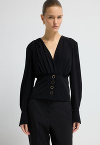 Blusa peplum nera con maniche lunghe, scollatura a V profonda, dettagli plissettati e bottoni dorati sul davanti. Tessuto morbido con una vestibilità strutturata.