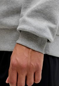 Lähikuva henkilön kädestä, jossa on harmaalla pitkähihainen huppari ja mustat housut, esittäen ihon tekstuurin ja kankaan yksityiskohdat.