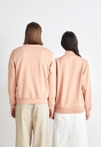 Felpe in cotone rosa viste di spalle, con polsini e orli a coste. I portatori le abbinano a pantaloni e gonne di colori chiari.