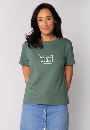 Groene katoenen t-shirt met een ronde hals en korte mouwen. Witte tekst in een speelse lettertype leest "Ich zähl bis drei!" op de voorkant.