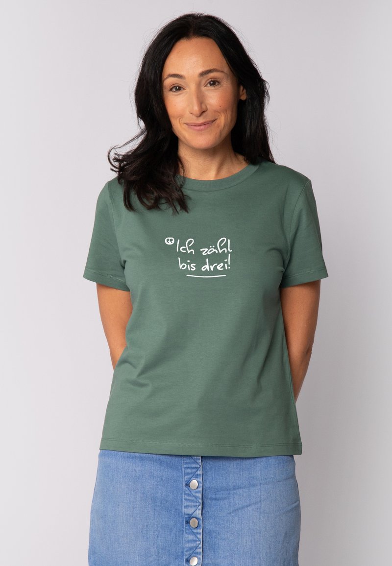 Grünes Baumwoll-T-Shirt mit rundem Halsausschnitt und kurzen Ärmeln. Auf der Vorderseite steht in verspielter Schrift "Ich zähl bis drei!" in weißer Farbe.