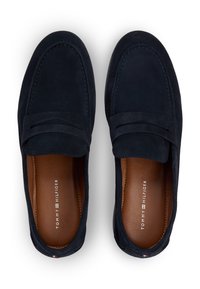 Μοκασίνια σε σκούρο μπλε σουέντ με ραμμένο σχέδιο μοκασίνου, διακοσμητικό λουράκι και καφέ δερμάτινη εσωτερική επένδυση. Το λογότυπο του κατασκευαστή είναι ορατό στο εσωτερικό.