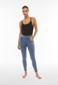 Nainen seisoo paljain jaloin musta hihaton toppi ja siniset istuvat jeggingsit, joissa on taskut, kädet rentoina ja yhdessä taskussa.