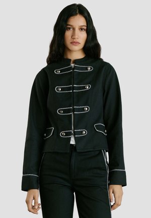 Femme aux longs cheveux foncés portant une veste militaire noire avec des garnitures et des boutons argentés, associée à un pantalon noir assorti avec des bandes blanches.