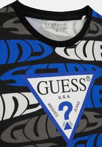 T-shirt en coton biologique noir avec des motifs graphiques bleus et gris, doté d'une étiquette triangulaire blanche affichant le logo et les détails "GUESS".