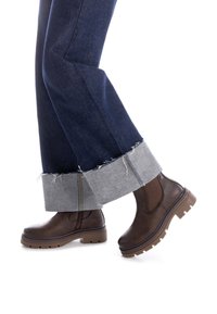 Botas de tobillo de cuero marrón con suela gruesa, combinadas con jeans azul oscuro que tienen un dobladillo de denim gris doblado con bordes deshilachados.