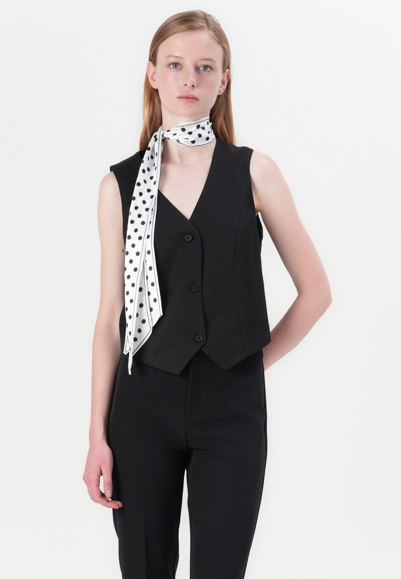 Gilet sans manches noir au design ajusté, doté de trois boutons, associé à une écharpe blanche ornée de pois noirs.