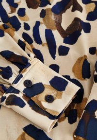 Tissu à motif léopard avec des couleurs bleu, marron et beige. Présente des poignets boutonnés et une texture douce et lisse.