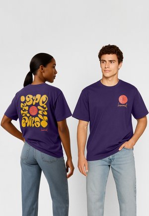 Twee personen dragen paarse t-shirts met oranje basketballogo's; de een toont de voorkant, de ander de achterkant met gestileerde gele tekst.