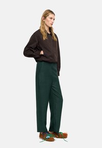 Brązowa kurtka na zamek, zielone teksturowane spodnie z wysokim stanem oraz brązowe buty z kolorowymi sznurówkami. Model stoi na tle jednolitego tła.