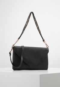 Černé kožené psaníčko s texturou a klopou ve tvaru obálky, odnímatelný černozlatý pletený řetízkový popruh, vystavené na bílém povrchu.