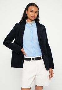 Mornarski sako od korduroja preko svijetloplavog kardigana na gumbe, upareno s bijelim kratkim hlačama i crnim remenom. Ima teksturiranu tkaninu i gumbe.