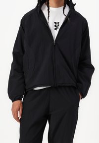 Chaqueta negra ligera con cremallera y puños y dobladillo elásticos, que presenta una textura sutil y bolsillos laterales, llevada sobre una camisa blanca.