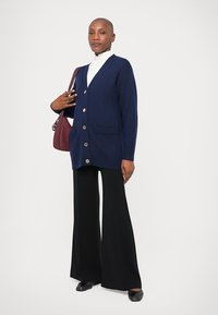 Σκούρο μπλε πουλόβερ με μεγάλους αιχμηρούς, δύο μπροστινές τσέπες, φορεμένο πάνω από λευκή ζιβάγκο, σε συνδυασμό με μαύρα φαρδιά παντελόνια και μαύρα παπούτσια.