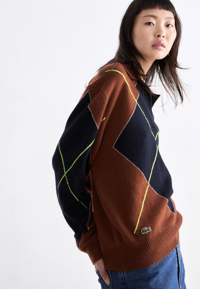 Pull marron et bleu marine avec motifs en losanges et accents néon ; fabriqué dans un matériau doux ; possède des poignets côtelés et un logo en bas.