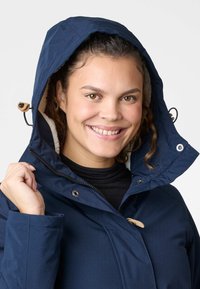 Granatowa wodoodporna kurtka z kapturem wyściełanym futrem i beżowymi akcentami, wyposażona w zamek błyskawiczny, zatrzaski oraz troczki do regulacji.