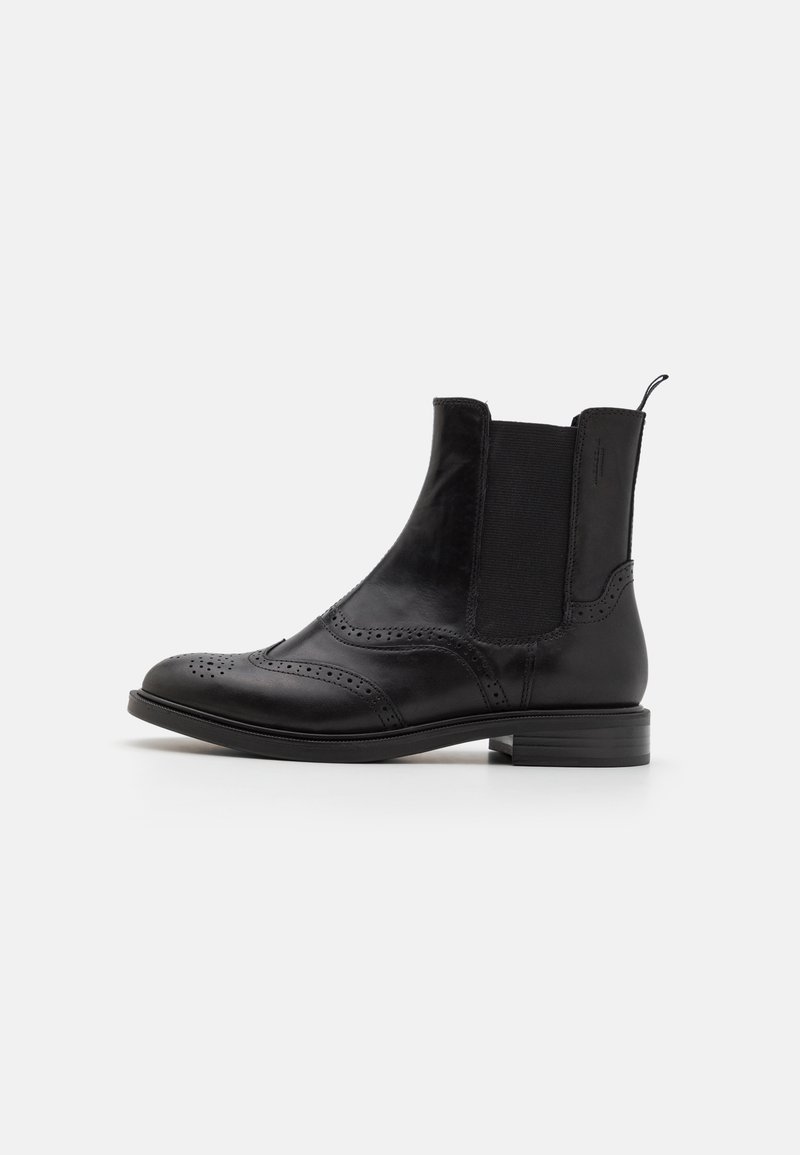 Bottine en cuir noir avec détails brogue, panneau latéral élastique et petit talon empilé sur fond blanc uni.
