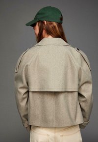 Jachetă din amestec de lână bej, cu un design scurt, epauleți pe umeri și două butoane, asortată cu o șapcă de baseball verde.
