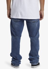 Vaqueros de mezclilla azul con un corte relajado, que presentan una textura desgastada, bolsillos traseros y detalle del logo. Combinados con una camiseta blanca y zapatillas negras.