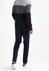 Homme portant un pull en tricot noir, gris et blanc, un pantalon bleu foncé et des baskets blanches, marchant loin sur un fond blanc.