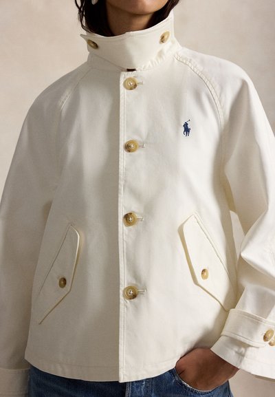 Λευκό μπουφάν με κουμπιά και ψηλό γιακά, δύο μπροστινές τσέπες με καπάκι, μικρό κέντημα με λογότυπο παίκτη πόλο σε ναυτικό μπλε χρώμα, φορεμένο από άτομο με χέρι στην τσέπη.