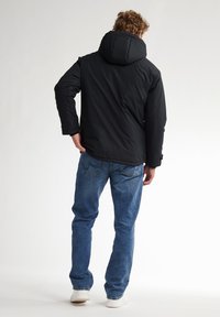 Giacca a vento nera con cappuccio, caratterizzata da una texture liscia e polsini elasticizzati, indossata sopra jeans blu e sneakers bianche. Vista posteriore.