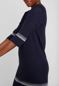 Una donna che indossa un vestito blu scuro con maniche a tre quarti, che presenta un motivo a righe vicino ai polsi, mentre posa su uno sfondo chiaro.