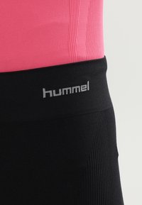 Gros plan sur une ceinture noire avec le logo gris "hummel" portée sur un tissu côtelé rose.