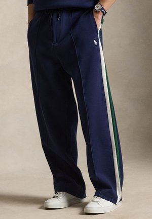 Osoba ubrana w granatowe spodnie dresowe z białymi i zielonymi paskami po bokach, białe sneakersy oraz zegarek na lewej ręce, z ręką w kieszeni.