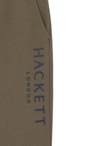 Maslinaste zelene trenerice izrađene od mekane tkanine, s vidljivim bočnim dŽepom i velikim plavim natpisom "HACKETT LONDON" duž noge.