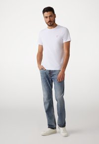 Witte katoenen t-shirt met een klein rood driehoekig logo, gecombineerd met lichtblauwe denim jeans en witte sneakers. Casual pasvorm, korte mouwen.
