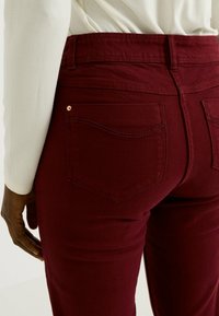 Jeans burdeos de cintura alta, con bolsillos traseros y detalles de tachuelas doradas. La tela parece suave, con una textura lisa y una forma ajustada.