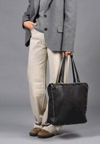 Zwarte leren tote bag met zilveren studs, rechthoekige vorm en stevige schouderbanden, gecombineerd met lichtbeige broek en grijze jas.