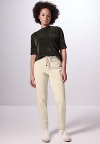 Diepgroene shirt met korte mouwen van textuurstof, gecombineerd met beige broek met trekkoord. Model draagt witte sneakers. Minimalistisch ontwerp.