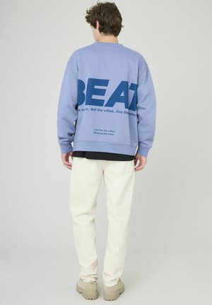 Sweatshirt bleu clair avec un grand texte bleu marine au dos, porté avec un pantalon blanc et des chaussures beiges, sur un fond uni.