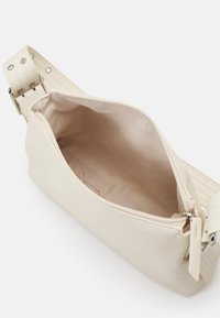 Sac bandoulière rectangulaire beige avec une large sangle réglable et une fermeture éclair ouverte montrant la doublure intérieure vide.