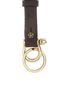 Fettuccia portachiavi in pelle marrone scuro con accessori in ottone, dotata di due anelli intrecciati e un dettaglio decorativo con bottone in rilievo.
