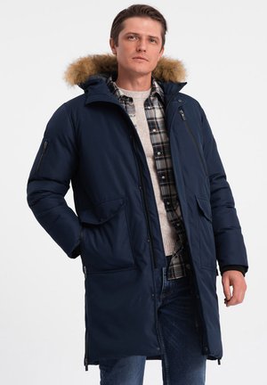 Marineblauer Daunenmantel mit gefütterter Kapuze aus Fell, zwei Seitentaschen und einer mit Reißverschluss versehenen Brusttasche. Besitzt gerippte Bündchen und ein mittellanges Design.