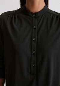 Chemise noire à boutons avec un col montant, comportant six boutons et des détails froncés aux épaules, fabriquée en tissu doux et extensible.