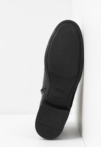 Semelle d'une chaussure formelle en cuir noir avec une semelle minimale, marquée "BOSS", exposée debout sur son talon contre un fond blanc d'angle.