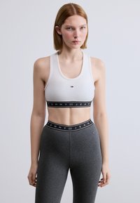 Λευκό αθλητικό σουτιέν με στρογγυλή λαιμόκοψη, διακοσμημένο με μαύρη ταινία που γράφει "TOMMY JEANS", συνδυασμένο με εφαρμοστά σκούρα γκρι κολάν.