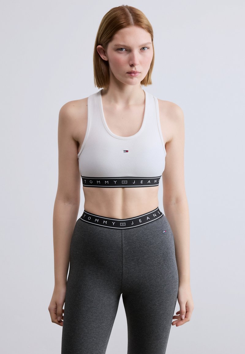 Λευκό αθλητικό σουτιέν με στρογγυλή λαιμόκοψη, διακοσμημένο με μαύρη ταινία που γράφει "TOMMY JEANS", συνδυασμένο με εφαρμοστά σκούρα γκρι κολάν.