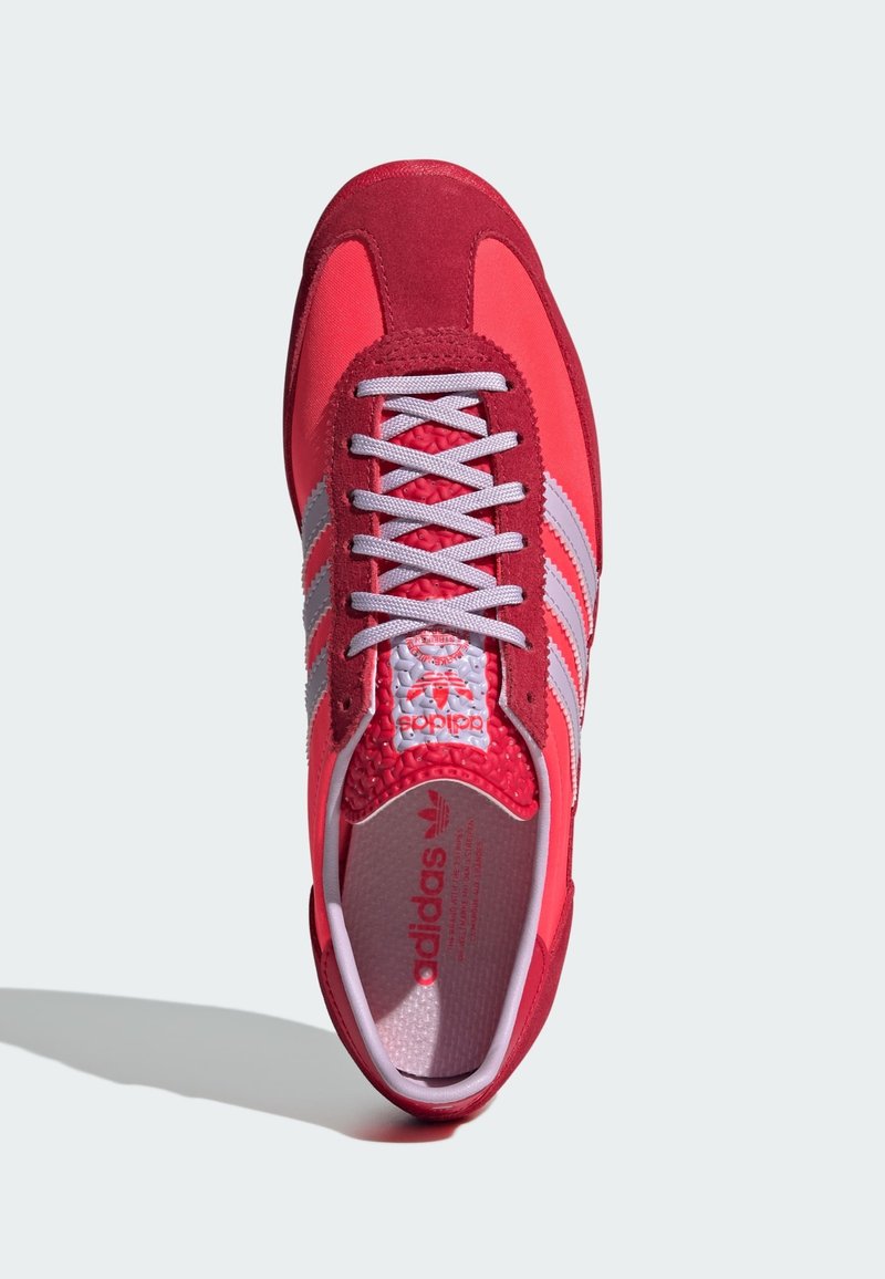 Zapatilla deportiva roja y rosa con materiales de ante y malla, que presenta tres rayas grises, lengua texturizada y cordones de color púrpura claro.