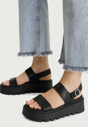 Feet wearing black platform sandals with two straps and silver buckle, paired with light blue frayed hem jeans.