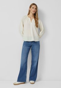 Blusa strutturata color crema con scollatura a V e dettagli arricciati, abbinata a jeans a gamba larga blu e sneakers chiare.