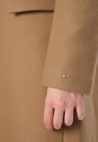Ruka blízko manžety rukáva z camel hnedého vlnenej kabátu so malou obdĺžnikovou červenou, bielou a modrou kovovou logom odznakom.
